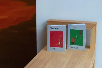 Two small booklets with red square image on one and a green square images on the other, standing up on the end of a light wooden bench in front of a white wall. The text on the covers read "Flare-Up Katie Kitamura in collaboration with Goldin+Senneby"