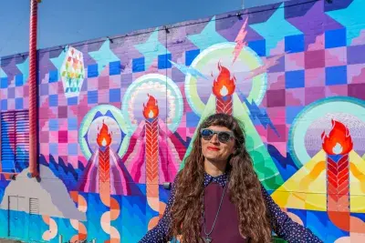 Portrait of a woman wearing sunglasses with long hair standing in front of a brightly colored patterned mural wall.