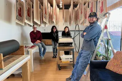 Four people standing around a printing press machine in a small room with a window on the right side. There are paper artworks hanging from the ceiling in a row.