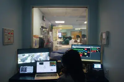 In a small dark control room, a person sits in front of multiple computer monitors while observing people in an adjoining patient room via an interior window.