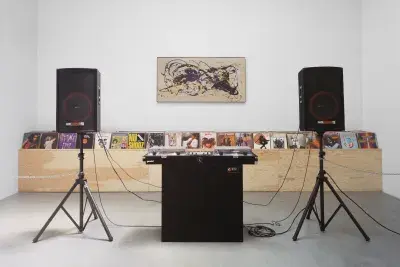 A record table with two record trays is in the middle of the room between two speakers. Wooden shelf holding records is against the white wall behind.