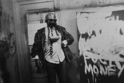 Black and white image of a person wearing a gas mask and helmet stands in a room with graffiti covering the walls.