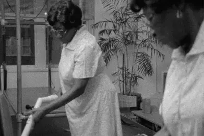 Black and white photograph of an older woman holding a PVC pipe like a vaccum cleaner. Her reflection can be seen in the mirror.