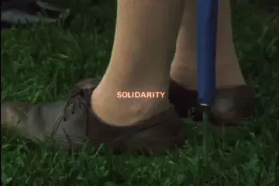 Two feet wearing leather lace up shoes stand in green grass with the bottom of a blue umbrella to the side. Orange text reads "SOLIDARITY"