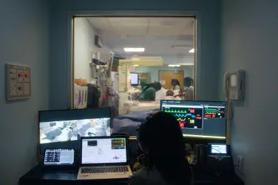 In a small dark control room, a person sits in front of multiple computer monitors while observing people in an adjoining patient room via an interior window.