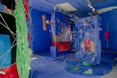 A room with two perpendicular walls covered with sculptures in Bright green and red pop against the royal blue space. Two sculptures that resembles shelled peanuts are attached to the center walls.