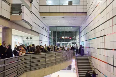 Group of people at an opening reception in an atrium. Stairs on the right hand side show one person walking on the basement level below.