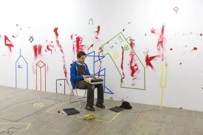A man sits on a folding chair and types on a typewriter. The white wall behind him and the wooden floor are smeared with red paint and marked with multicolored tape in various geometric forms. Other objects are scattered at his feet, including a corded phone, a folder, and a hat.