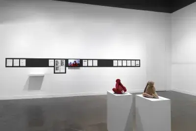 In the foreground, two bunny sculptures—one red, one beige—sit on pedestals, facing away from each other. In the background, on the wall, a stripe of black paint outlines documents tacked in a row (some of their contents redacted in black rectangles) and a video monitor showing a woman’s head. On the far right, a different wall is marked by flowing horizontal lines and a drippy substance.