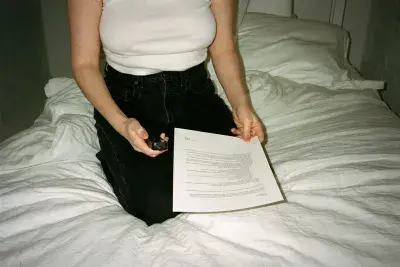 A woman kneels on a bed with white covers, holding an audio recorder in one hand and a printed textual score in the other.