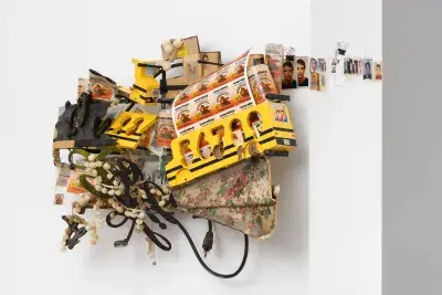 A wall-mounted assemblage sculpture featuring yellow toy buses with 'Wardley' stickers, black electrical cords, and white beads. Small photographs are integrated into the sculpture and extend horizontally to the right. A floral-patterned fabric forms the base of the dense arrangement.