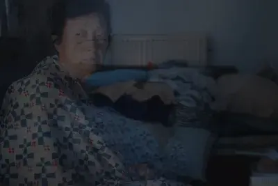 A translucent image shows an older person, wrapped in a blue, white, and red patterned blanket, looking forward with a neutral expression. The dimly lit background reveals a room with a radiator and piled blankets.