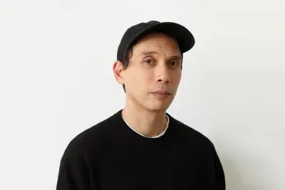 Man in a black long sleeve shirt and a black baseball cap is sitting looking directly at the camera in front of a beige wall.