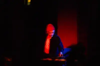 Man with a mustache wearing a beanie and a dark jacket playing a dj controller drenched in red light.