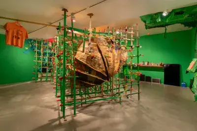 A large, textured, brown cardboard sculpture, is encased within a green wooden grid structure. The installation is set in a room with bright green walls, and an orange t-shirt hangs from the ceiling on the left side of the sculpture.