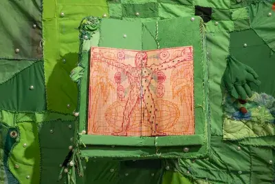 An open wooden book with a pixelated illustration of a human figure in orange, pink, and red is centered on a raised section of green fabric. The fabric is part of a larger green patchwork adorned with white pearls, and a green fabric hand emerges from the right, in front of a patch of colorful textured material.