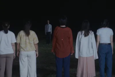 Video still of a group of people standing outdoors at night, seen mostly from behind, facing a person who stands apart from the group.