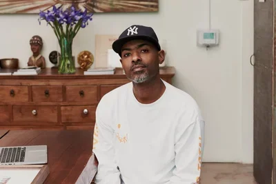 Man wearing a white longe sleeve shirt and a black baseball hat is sitting in a living room space with a wood side table behind with purple flowers in a vase sitting on the table.