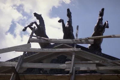 Video still showing a building rooftop adorned with three sculptural horses hanging off the front as if they are mid-gallop.