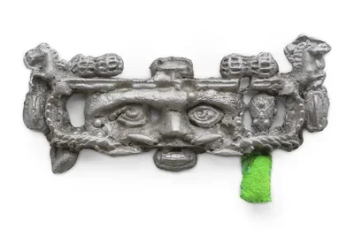 A sculpture made of gray aluminum shaped into a long, horizontal, decorative shape with intricate reliefs and raised figures. The piece includes carved eyes with peanut and horse motifs, and a bright green fabric element attached to the right underside, adding a pop of color.
