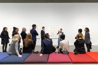 A group of people are standing around and seated on a colorful bench in a gallery as artist Christine Sun Kim talks near her work.