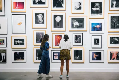 Two women wearing colorful clothing stand in the List Center galleries during the Student Lending Art Program 2019 and look at works of art on the walls.