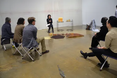 Guests are seated on small stools around a woman with dark hair and glasses who is leading the lecture inside the gallery space.