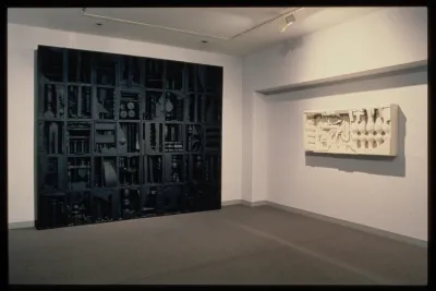 A sculptural wall of wood pieces painted black rests against a wall. A white wood sculpture hangs on the opposite wall. 