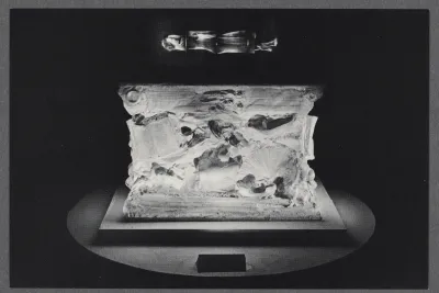 A black and white photo of a ceramic sculpture. The sculpture is a cube shape with faded details, spotlit on a pedestal.
