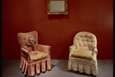 Two decorative chairs set in front of a red wall with a gold framed mirror hung between them.