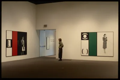 A viewer stands by the entrance to the gallery between two works, both containing figures, solid colors, and graphic symbols. 