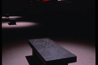 Sign reading “If you aren’t political your personal life should be exemplary” hung in a dark room with three benches spotlit.