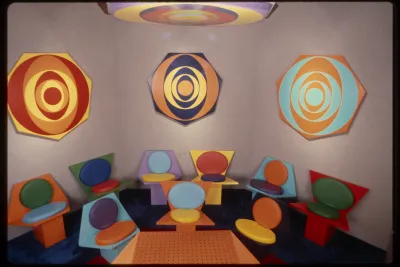 Rainbow target on ceiling above three heptogonal shapes with colorful concentric circles hanging above 2 rows of colorful seats.