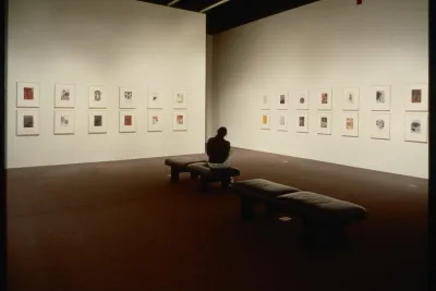 A visitor sits on gallery benches while viewing the framed works that are hung on the walls around them.  
