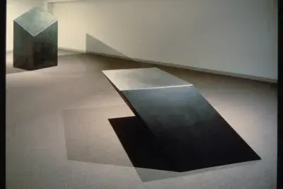 Two large, black, metal, geometric sculptures sit in a gallery.