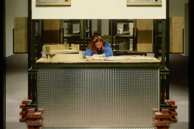 A visitor sits inside a cubicle made from industrial materials while reading a newspaper.