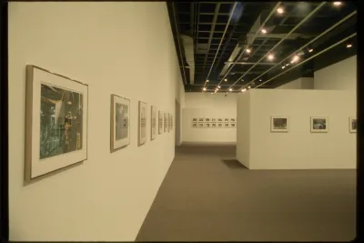 Eleven small framed photographs hang in the middle of gallery walls. Fourteen small photographs hang in the distance.