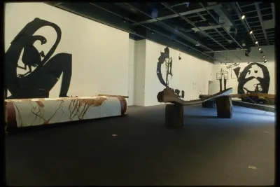 Gallery view of four giant black paper cut outs of abstract figures on the walls, and three wooden slabs and logs on the floor.