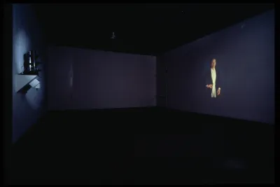 A dark gallery space shows a projected film. In the still there is a figure posing with their hand on their hip.