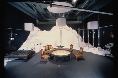 An installation of a room with a table, chairs, and bed. These elements sit before a constructed white mountain.