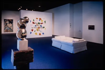 An abstract sculpture stands in the foreground while a white couch is settled behind. Hanging works are on the surrounding walls.