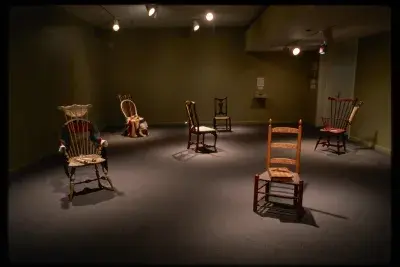 Dim room with spotlights on six spaced out colorful 17th and 18th century chairs with household items sitting on them. 