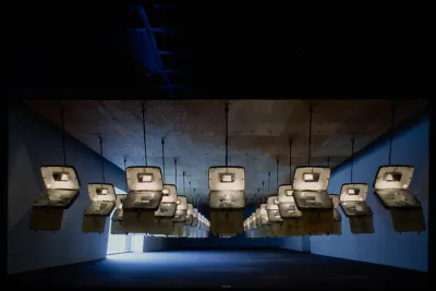 Identical opened suitcases are hung from the ceiling in rows. Each has text projected on the inside.