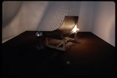 A wire mesh sculpture of a reclined chair on a wooden base. A light shines through, projecting a fence pattern on the wall. 