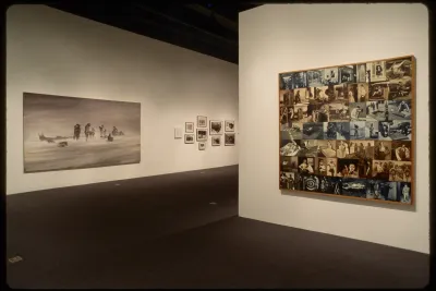 Large black and white oil painting of desert nomads and ten framed photos hang on adjacent wall to patchwork of sepia photos.