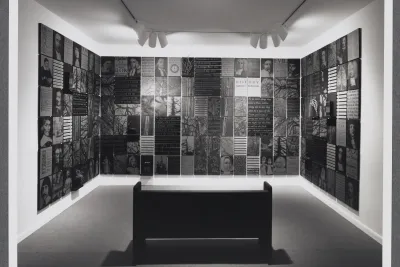 A bench sits in front of a gallery wall. On the wall are photos of historical portraits as well as historical documents.  