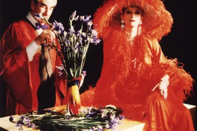 A man and a woman arer dressed in orange. The woman on the right wears a furry coat and a large brimmed hat with orange fringe.
