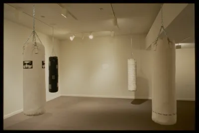 Three white and one black punching bags with text and images printed on them hang in four corners of gallery. 