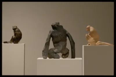 Three abstracted clay sculptures of apes sit on white gallery pedestals. From left to right: Brown and black, black and grey, tan. 