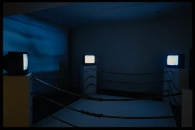 A boxing ring where the corners are made from pedestals with monitors. The blue light from the monitors shine on the walls.